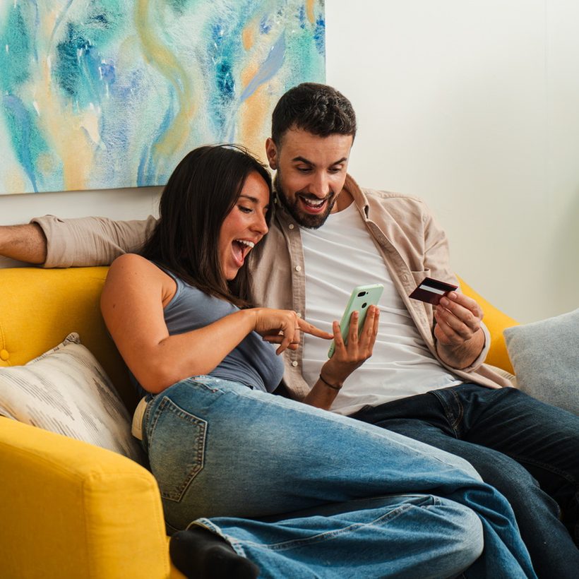 A couple on a sofa smiling at a phone, one holding a bank card